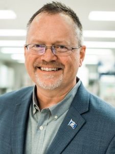 This is a headshot of Brent Sanford. He is wearing glasses, a gray shirt and gray suit jacket; the background appears to be a brightly lit office space. He is smiling directly at the camera.