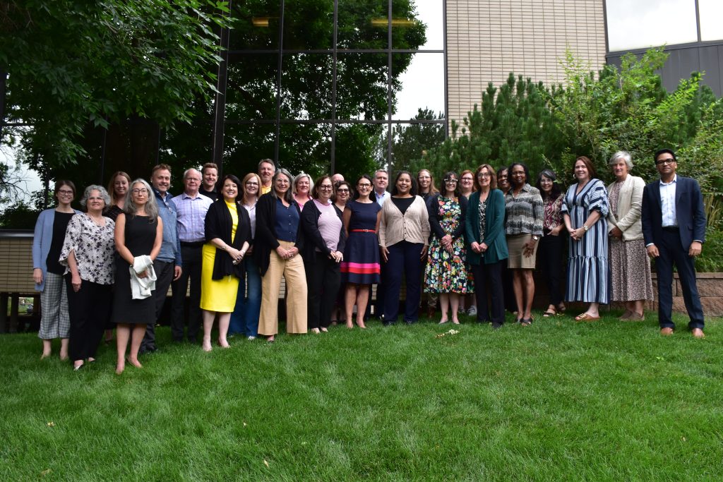 This is a group photo of 19 people smiling at the camera and standing outside on the grass before an office building. They are wearing assorted business attire. They are participants in the 2025 Academy hosted by WICHE.