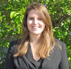 A woman with long brown hair faces the camera and is smiling. She is wearing a black top and black suit jacket. She is outside in front of greenery. 